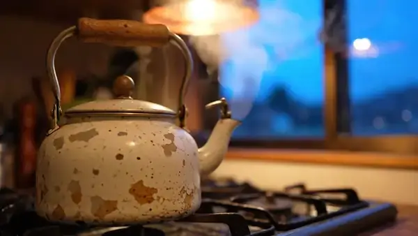 Steam curling from the spout of an enamel kettle on a stovetop as the kitchen window darkens